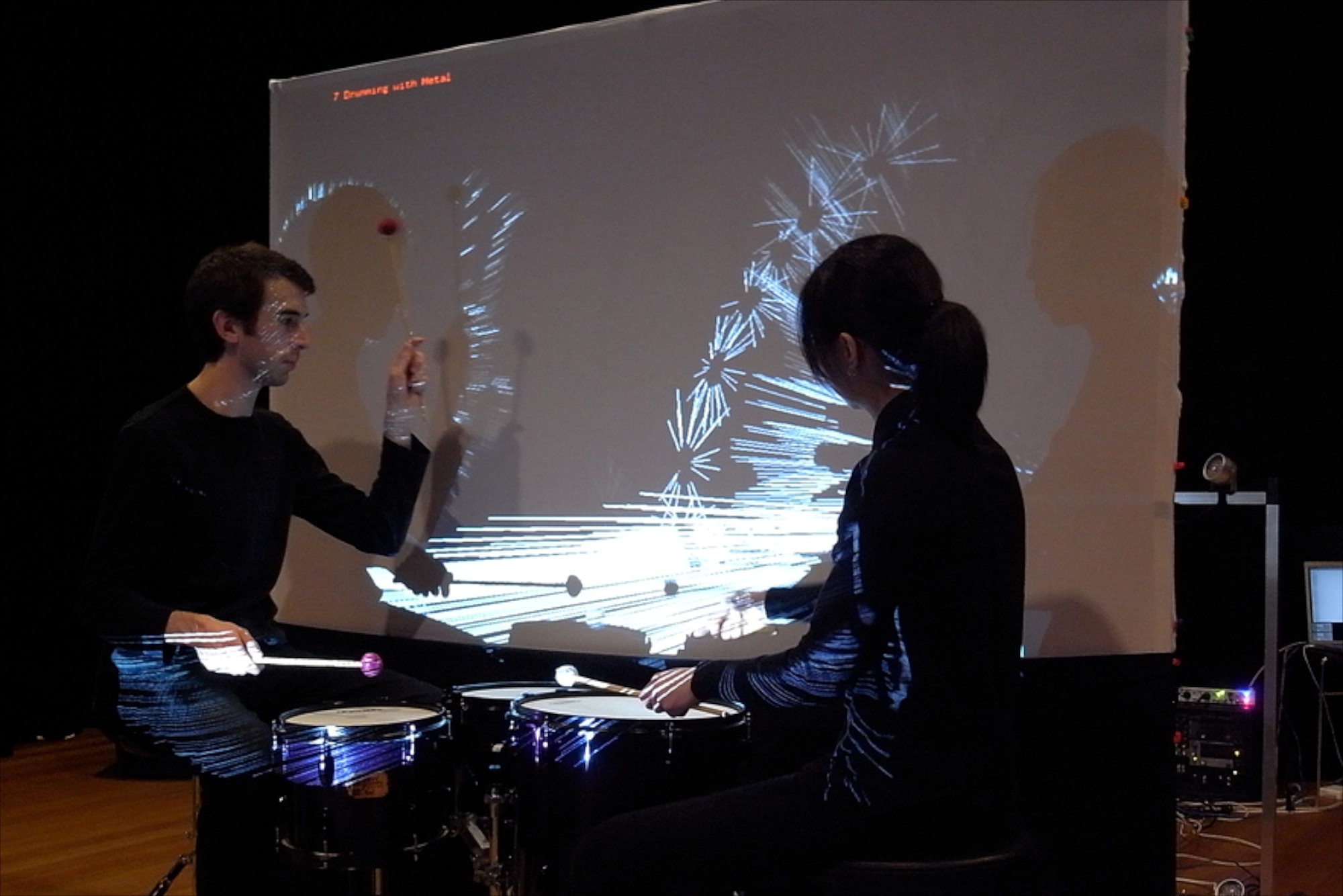 Charles Martin and Chi-Hsia Lai performing Strike on Stage. The performers sit in front of the screen and move sticks to trigger sounds.