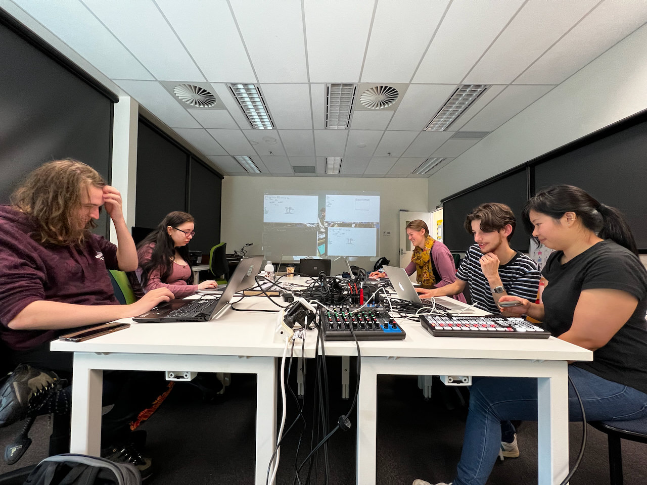 Students in a laptop ensemble group session, gathered around laptops and a mixer in 2022